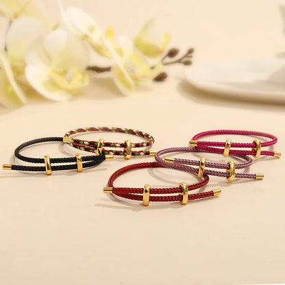 Set of four braided bracelets with gold accents on a beige surface with blurred flowers in the background